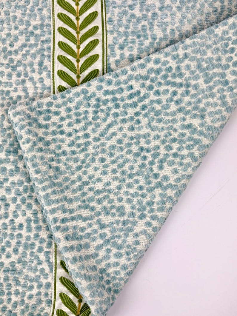 May include: Close-up of a textured fabric with a repeating pattern of small, light blue dots on a white background. A folded section reveals the fabric's texture. A vertical stripe with green leaf designs on a white background adds a decorative element.
