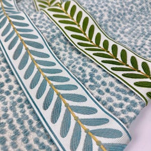 May include: Close-up of two decorative ribbons with leaf patterns. One ribbon features blue leaves with a gold stem, the other has green leaves with a gold stem. Both ribbons are on a textured blue and white fabric.