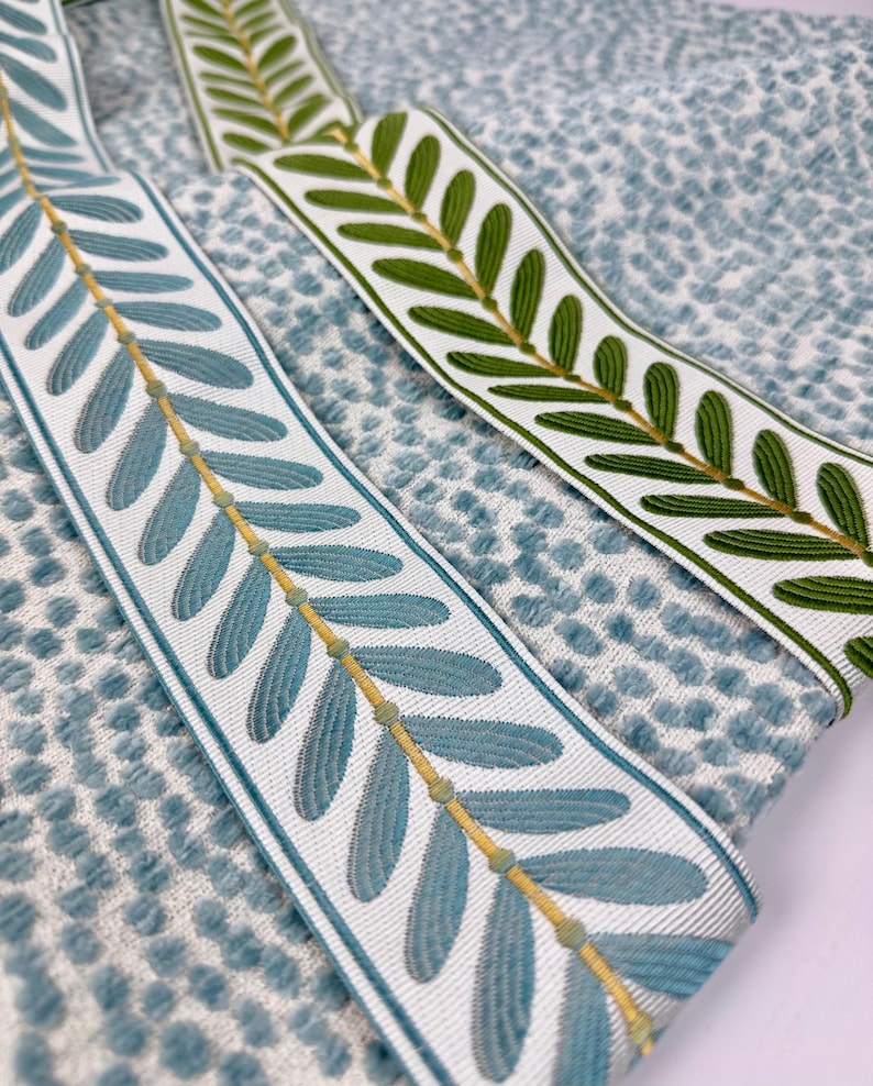 May include: Close-up of two decorative ribbons with leaf patterns. One ribbon features blue leaves with a gold stem, the other has green leaves with a gold stem. Both ribbons are on a textured blue and white fabric.
