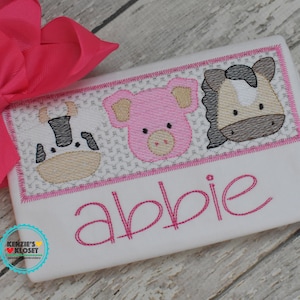 May include: White onesie with pink and brown embroidered farm animals, including a cow, pig, and horse. The onesie has the name "Abbie" embroidered in pink below the animals.