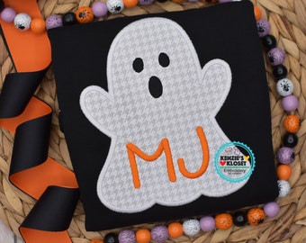 May include: A black t-shirt with a white ghost applique with a houndstooth pattern. The ghost has orange eyes and a mouth. The letters "MJ" are embroidered in orange inside the ghost. The t-shirt is on a woven wicker surface with a purple and orange beaded necklace.