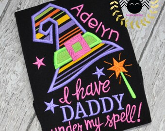 May include: Black t-shirt with a colorful embroidered witch hat, stars, and a magic wand. The text reads "Adelyn" and "I have DADDY under my spell!" in pink and purple thread. A Halloween-themed design.