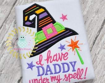 May include: White t-shirt with an embroidered witch hat, stars, and a magic wand. The text reads "I have Daddy under my spell!"
