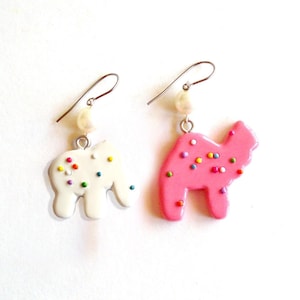 May include: A pair of dangle earrings featuring a pink and white frosted sugar cookie shaped like a camel. Each earring has a pearl charm and a silver hook.