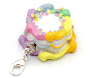 May include: A colorful cake charm with a silver lobster claw clasp. The cake is decorated with pastel pink, yellow, blue, and green frosting and sprinkles.