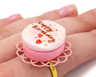 May include: A novelty ring shaped like a pink birthday cake with white frosting, sprinkles, and the words "Happy Birthday." The ring has a gold band and a pink, lacy base.