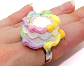 May include: A pastel-colored cake ring with yellow, pink, and purple frosting. The ring is made of metal and has a white base.