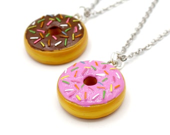 Könnte beinhalten: Zwei silberne Kettenhalsbänder mit Donut-Anhängern. Ein Donut ist rosa mit bunten Streuseln und der andere ist braun mit weißen Streuseln.