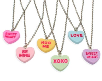 Op de afbeelding: Een verzameling hartvormige hangerkettingen in verschillende pastelkleuren, elk met een andere boodschap in roze tekst. De kettingen hebben zilverkleurige kettingen. Berichten zijn onder meer "SWEET HEART", "BE MINE", "HUG ME", "XOXO" en "LOVE".