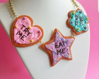 Può includere: Una collana stravagante con ciondoli a forma di biscotto. Il ciondolo a forma di cuore ha il testo "TRY ME", il ciondolo a forma di stella ha il testo "EAT ME" e il ciondolo a forma di fiore ha il testo "TAKE ONE". La catena è dorata.