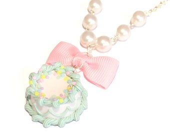 Puede incluir: Un dije de pastel verde pastel y blanco con chispas de colores, unido a un collar de perlas con un lazo rosa.