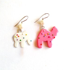 Pu&ograve; includere: Un paio di orecchini pendenti con un ciondolo a forma di biscotto rosa e bianco. Il ciondolo rosa ha la forma di un cammello e il ciondolo bianco ha la forma di un elefante. Entrambi i ciondoli hanno spolverate colorate.