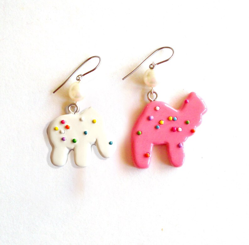 Pu&ograve; includere: Un paio di orecchini pendenti con un ciondolo a forma di biscotto rosa e bianco. Il ciondolo rosa ha la forma di un cammello e il ciondolo bianco ha la forma di un elefante. Entrambi i ciondoli hanno spolverate colorate.