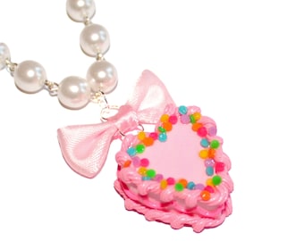 May include: A heart-shaped pendant necklace with a pink cake design. The cake is decorated with colorful sprinkles and pink frosting. A pink bow and a strand of white pearls complete the necklace.