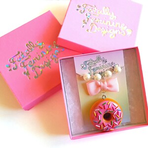 May include: A pink box with the text "Fatally Feminine Designs" in iridescent lettering. Inside the box is a donut-shaped necklace with a pink bow and white pearl accents. The necklace is on a white card with the same text as the box.