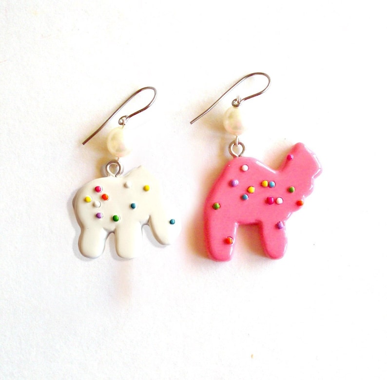May include: A pair of dangle earrings featuring a pink and white frosted sugar cookie shaped like a camel. Each earring has a pearl charm and a silver hook.