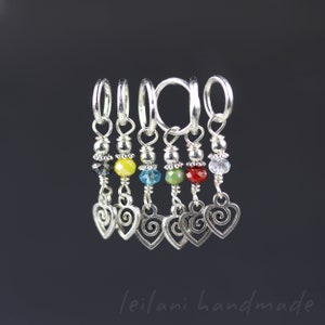 Peut inclure: Ensemble de sept marqueurs de point argentés. Chaque marqueur présente une perle de couleur différente et un charm en forme de cœur pendant. Les perles sont noires, jaunes, bleues, vertes, rouges et transparentes. Les breloques ont une forme de cœur avec un motif tourbillon.