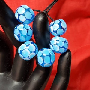 May include: A bracelet made of blue and white glass beads with a white polka dot pattern. The bracelet is tied with a black cord.