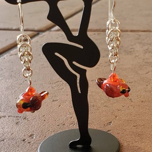 May include: A pair of silver earrings with a red and orange glass bird charm hanging from each. The earrings are on a black display stand.