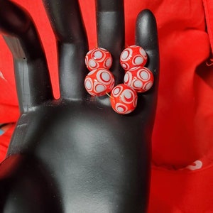 May include: Five red glass beads with white circles on them. The beads are in a hand.
