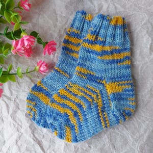Könnte beinhalten: Ein Paar handgestrickte Socken mit einem melierten Muster aus hellblauem, dunkelblauem und goldgelbem Garn. Die Socken haben ein geripptes Bündchen am oberen Rand und eine klassische Fersenkonstruktion mit einer gleichmäßigen Strickstruktur.