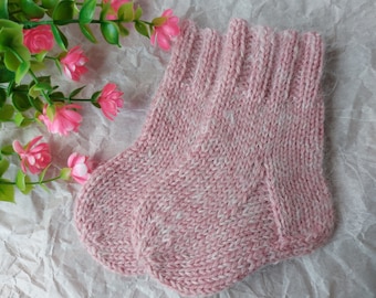 May include: A pair of soft, hand-knitted baby socks in a dusty rose pink colour. The socks feature a ribbed cuff at the top and a classic stockinette stitch pattern throughout, crafted from a fuzzy, textured yarn with subtle white flecks.