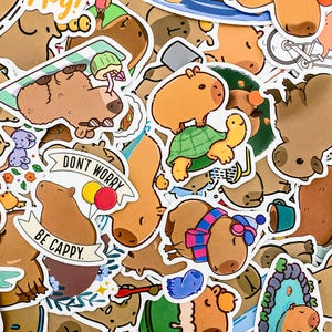 May include: A collection of colorful stickers featuring capybaras in various poses and settings. The stickers include phrases like "Don't Worry, Be Cappy" and depict capybaras riding turtles, wearing hats, and relaxing in nature.