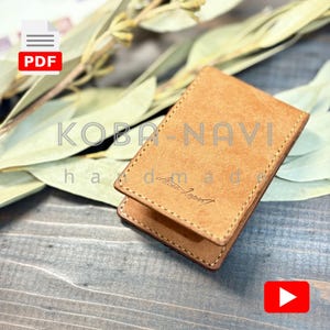 以下が含まれることがあります： ステッチされたエッジを持つ長方形の、明るい茶色の革製カードホルダー。「KOBA-NAVI」と「handmade」の文字が見えます。ホルダーは、乾燥した葉のある木製の表面にあります。