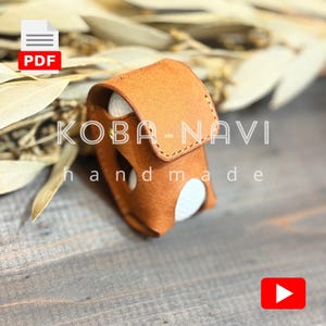 May include: A handmade, tan leather golf ball holder with a flap closure. The holder has a stitched design and holds a white golf ball. The text "KOBA-NAVI handmade" is visible on the holder.