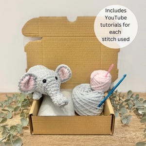 May include: A grey crocheted elephant sits in a cardboard box with grey and pink yarn, a crochet hook, and a yarn needle. The box includes YouTube tutorials for each stitch used. The elephant has pink inner ears.