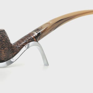 May include: A brown tobacco pipe with a textured bowl and a curved, light brown stem. The pipe rests on a silver-colored stand, showcasing its elegant design. The pipe is a classic accessory.