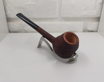 May include: A brown tobacco pipe with a textured bowl and a black stem, resting on a silver-colored pipe stand. The pipe has a classic design, suitable for smoking tobacco. The pipe is made of wood.