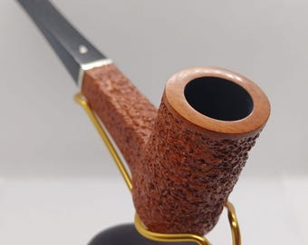 Puede incluir: Una pipa marrón y negra con un acabado texturizado, apoyada en un soporte negro con un soporte de metal dorado.