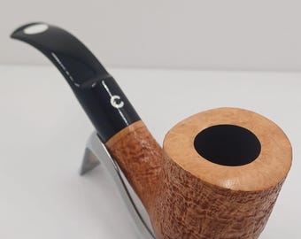 May include: A tobacco pipe with a light brown, textured bowl and a black stem. The pipe rests on a silver-coloured stand. The stem has a white accent. The pipe is designed for smoking tobacco.