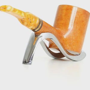 May include: A tobacco pipe with a light brown bowl and a yellow and brown stem. The pipe rests on a silver-colored stand with a curved design. The pipe is angled upwards, showcasing its polished finish.