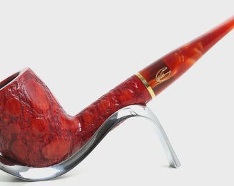 May include: A red tobacco pipe with a textured bowl and a smooth, marbled stem featuring a gold-coloured band and a small logo. The pipe rests on a curved, silver-coloured metal stand, showcasing a contrast between the rough bowl and stem.