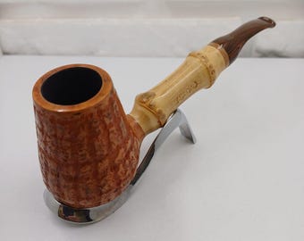 May include: A tobacco pipe with a textured brown bowl and a bamboo-style stem. The stem is light brown with dark brown accents. The pipe rests on a silver-coloured stand. The bowl is approximately 5 cm in diameter.