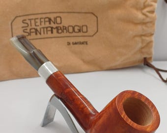 May include: A polished, reddish-brown tobacco pipe on a silver stand. The pipe has a silver band and a light-coloured mouthpiece. A tan drawstring bag with the text "STEFANO SANTAMBROGIO" is visible in the background.