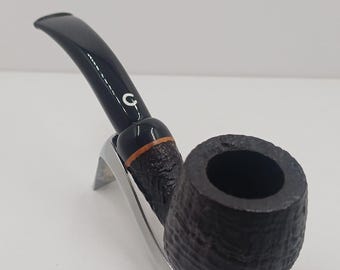May include: A dark brown tobacco pipe with a textured bowl and a black stem. The pipe rests on a silver-coloured stand. The stem has a white "G" logo. The pipe is designed for smoking tobacco.