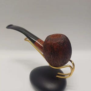 May include: A brown wooden pipe with a black stem resting on a gold metal stand. The pipe is on a black surface.