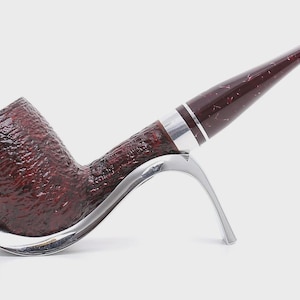 May include: A tobacco pipe with a dark red, textured bowl and a matching stem. The stem has a silver band near the bowl. The pipe rests on a silver-coloured stand, showcasing its elegant design.