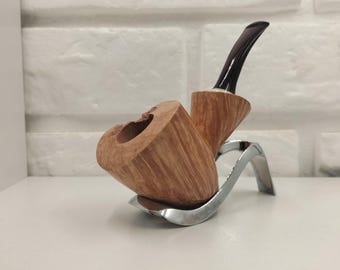 May include: A tobacco pipe featuring a light brown, natural wood grain bowl with a textured, organic finish. The pipe has a dark brown, curved acrylic stem and is resting on a polished, silver-colored metal stand with a curved, modern design.