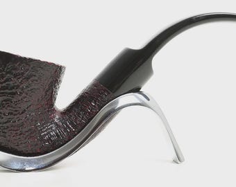 May include: A tobacco pipe with a dark brown, textured bowl and a smooth, black, curved stem. The pipe rests on a polished, silver-coloured metal stand that features a curved, open design to support the bowl and the stem of the pipe.