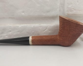 May include: A tobacco pipe featuring a textured, light brown bowl with a wide, angled rim. The shank is smooth and light brown, connected to a black, tapered stem by a thin, white, ring-shaped band. The pipe is positioned horizontally.