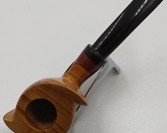 May include: A wooden tobacco pipe with a curved, light brown bowl and a black stem. The pipe features a decorative band with red and brown accents. The pipe is resting on a silver-coloured stand, showcasing its unique design and craftsmanship.