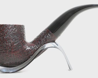 May include: A tobacco pipe with a dark brown, textured bowl and a smooth, curved black stem. The pipe is resting on a polished, silver-coloured metal stand that features a curved, open design to support the bowl and the shank.