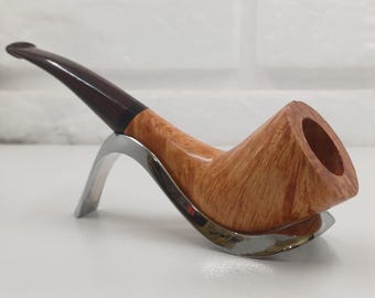 May include: A wooden tobacco pipe with a curved stem and a polished finish. The pipe is light brown with a dark brown stem and is resting on a silver-colored stand. The pipe has a classic shape and design.