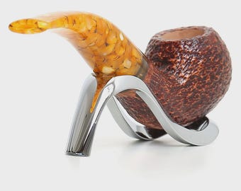 May include: A tobacco pipe featuring a dark brown, textured bowl and a translucent, amber-coloured stem with internal flecks. The pipe rests securely on a polished, silver-toned metal stand with a curved, modern design that supports the bowl.