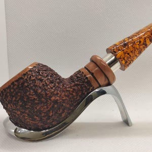 Könnte beinhalten: Eine Tabakpfeife mit einer dunkelbraunen, strukturierten Pfeifenschale und einem hellbraunen, gemusterten Mundstück. Die Pfeife steht auf einem silberfarbenen Ständer. Das Mundstück hat eine gesprenkelte, bernsteinfarbene Oberfläche.