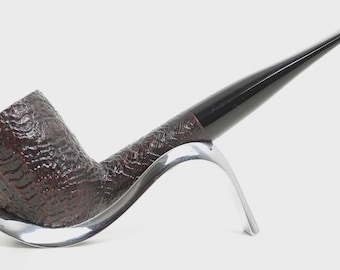 May include: A tobacco pipe with a dark brown, deeply textured bowl and a long, smooth, black stem. The pipe rests securely in a polished, silver-coloured metal stand that features a curved, modern design supporting the bowl and shank.